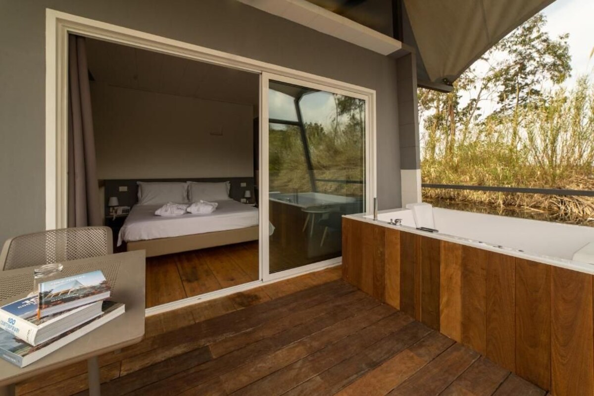 Ein modernes Schlafzimmer mit einer großen Schiebetür, die auf einen Balkon führt, Holzboden, einem Bett mit weißen Bettwäsche und einem kleinen Tisch mit einem Buch.