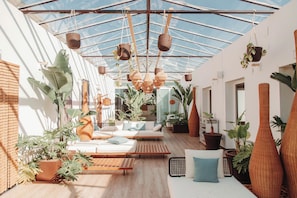 Un intérieur lumineux, baigné de soleil, avec un plafond en verre, des meubles en bois et diverses plantes en pot.