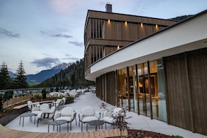 A modern building with a curved exterior, large windows, and a patio area with outdoor seating. - Ganischgerhof - Mountain Resort & Spa (Nova Ponente)