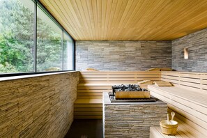 Eine moderne Sauna mit Holzbänken und einer Steinwand, große Fenster mit Blick auf einen Wald.