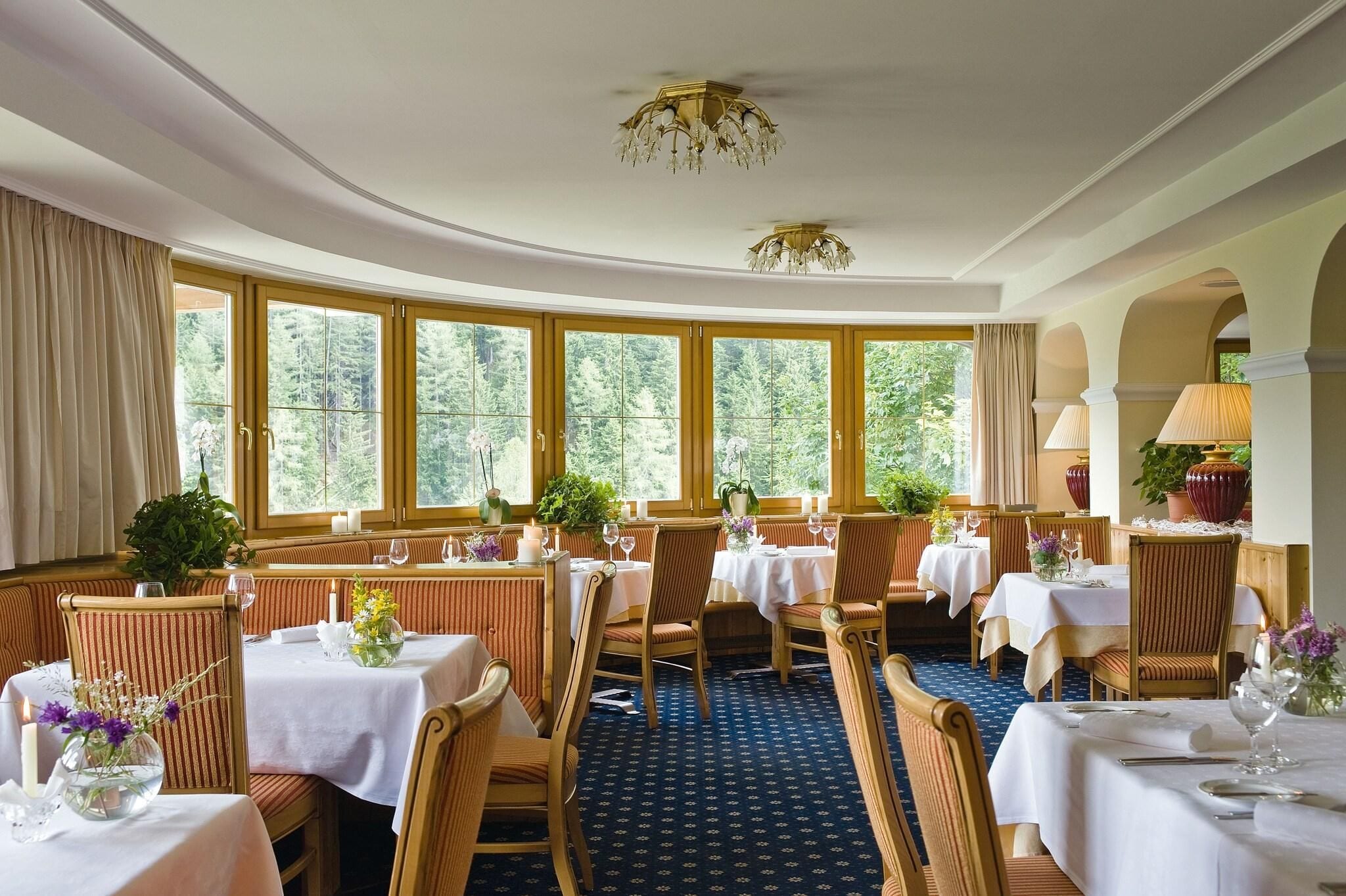 Un restaurante con mesas redondas, sillas y ventanas grandes con cortinas.