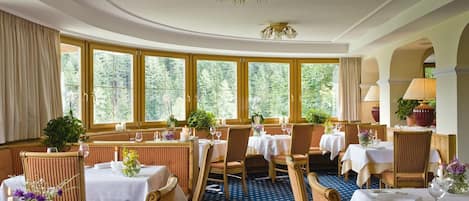 Un restaurante con mesas redondas, sillas y ventanas grandes con cortinas.