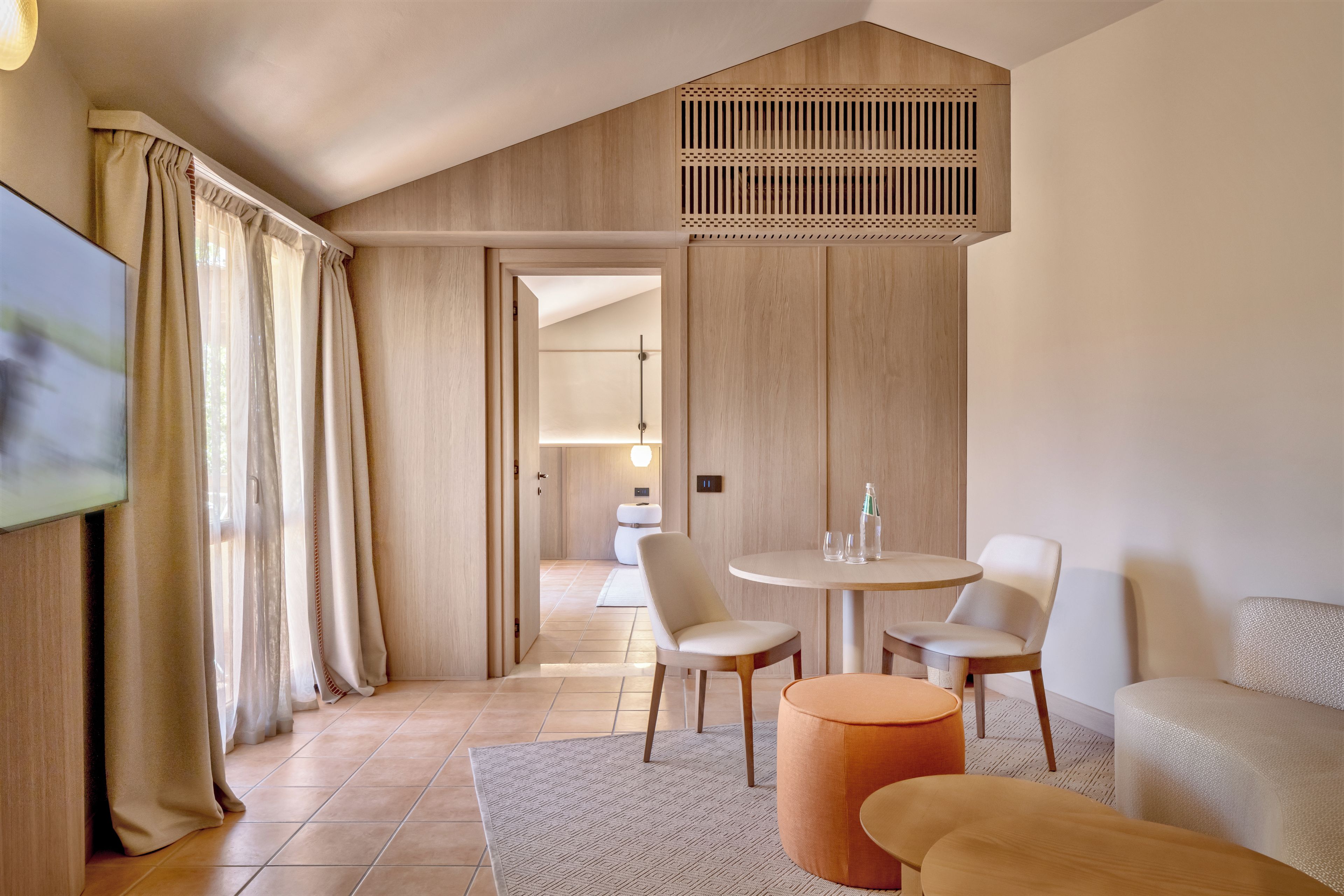 Una habitación de hotel moderna con techo de madera, un área de comedor con dos sillas y una mesa, un sofá y un televisor en la pared izquierda.