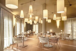 A spacious dining area with multiple round tables and chairs, large hanging pendant lights, and large windows with sheer curtains. - Veridia Resort, a Member of Radisson Individuals (Domus de Maria)