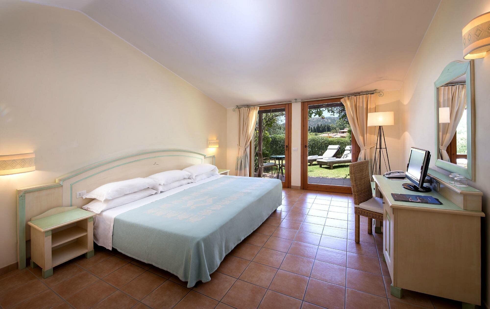 A bedroom with a bed, bedside tables, a desk with a computer, and a view of the outdoors.