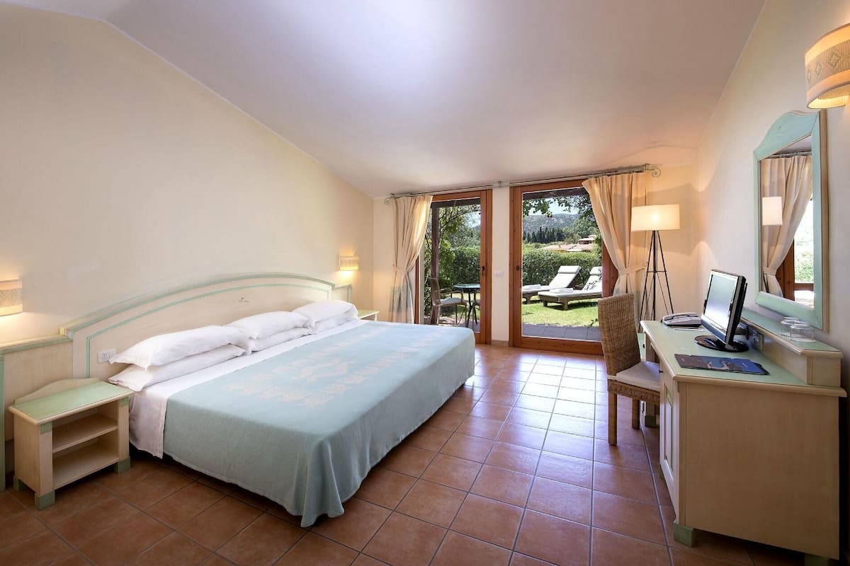 Quarto com cama, mesas de cabeceira, escrivaninha com computador e vista para o exterior.