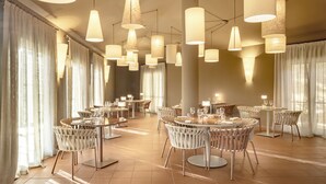 Un vaste espace dédié à la salle à manger, avec de nombreuses tables et chaises rondes, de grandes suspensions lumineuses et de grandes fenêtres ornées de rideaux légers.