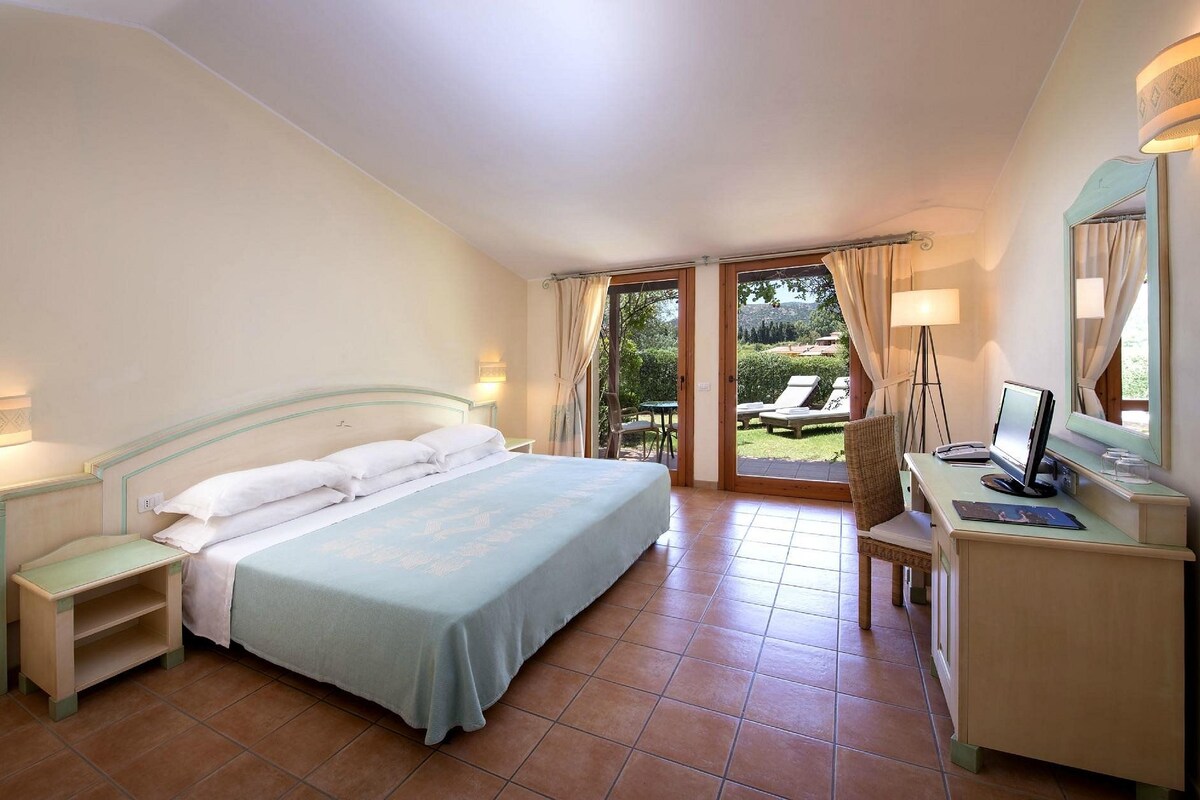 Un dormitorio con cama, mesitas de noche, un escritorio con computadora y vistas al exterior.