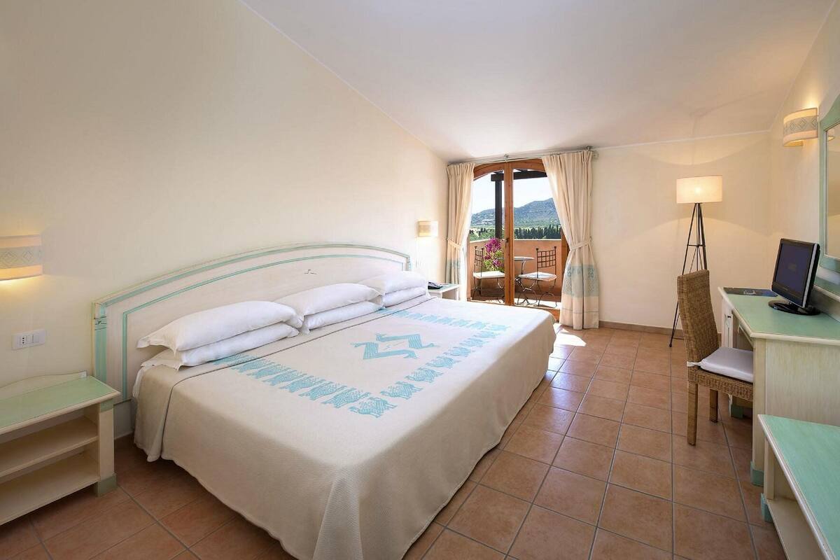Quarto de hotel com cama, escrivaninha com computador, cadeira e varanda com vista.