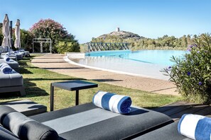 Zona de piscina con sillones de descanso y una pequeña mesa, rodeada de vegetación y vistas a una colina con una torre.