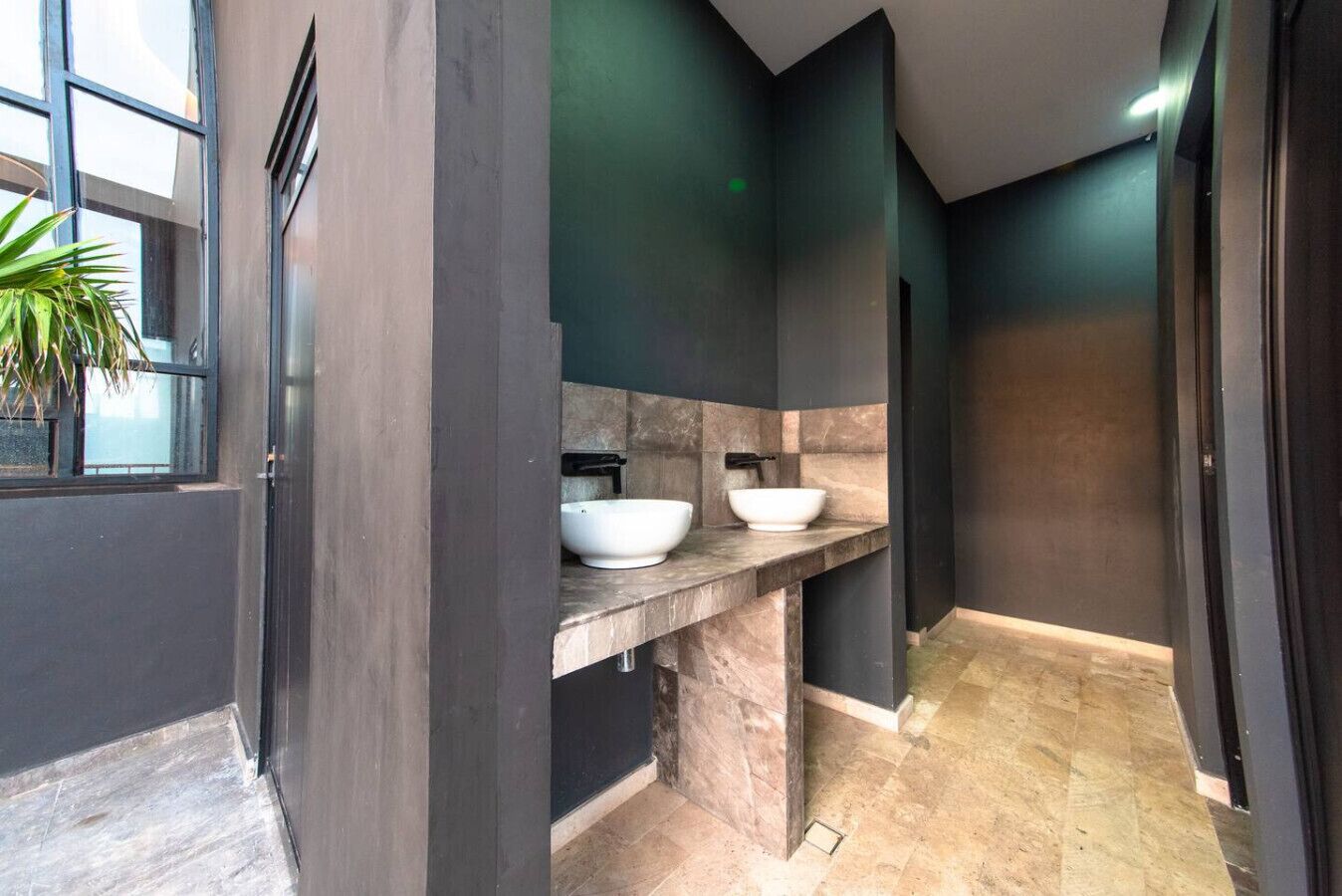 A modern bathroom with dark walls, two white vessel sinks, and a large window with a plant.