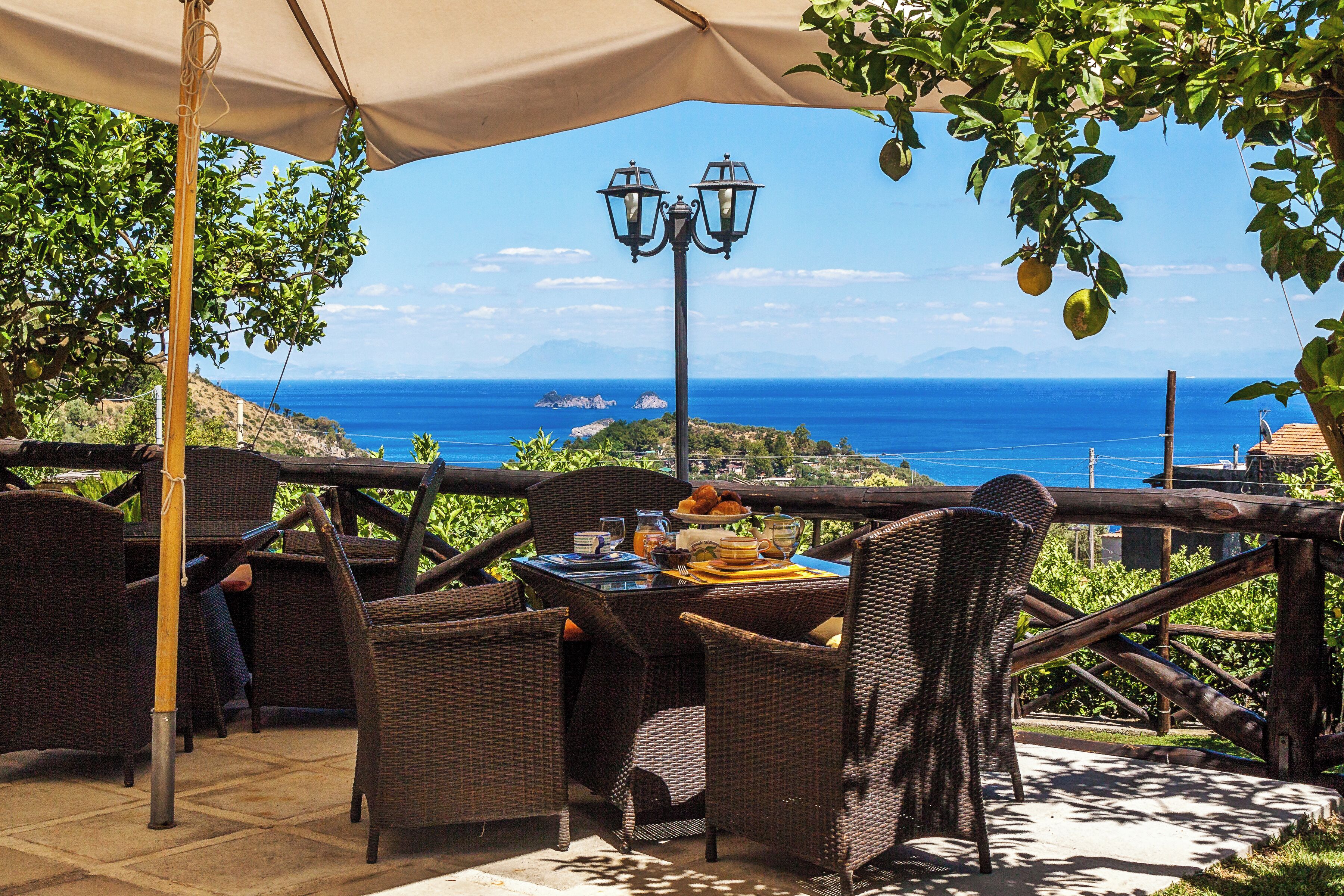 Uma área de refeições ao ar livre com mobiliário de vime, uma mesa posta com comida e bebidas, e vista para o mar.