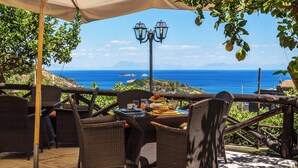 Uma área de refeições ao ar livre com mobiliário de vime, uma mesa posta com comida e bebidas, e vista para o mar.