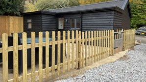 A small, dark-colored wooden cabin with a metal roof, surrounded by a wooden fence and gravel ground.