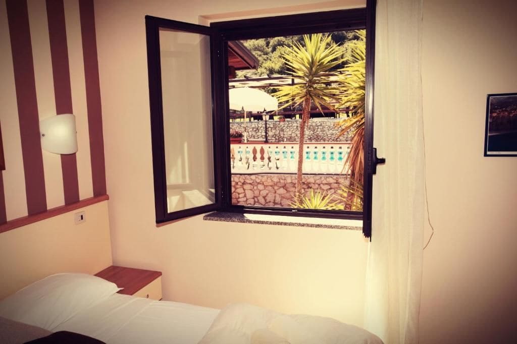 A bedroom with a bed, a window with a view of a pool and palm trees, and a wall-mounted light fixture.