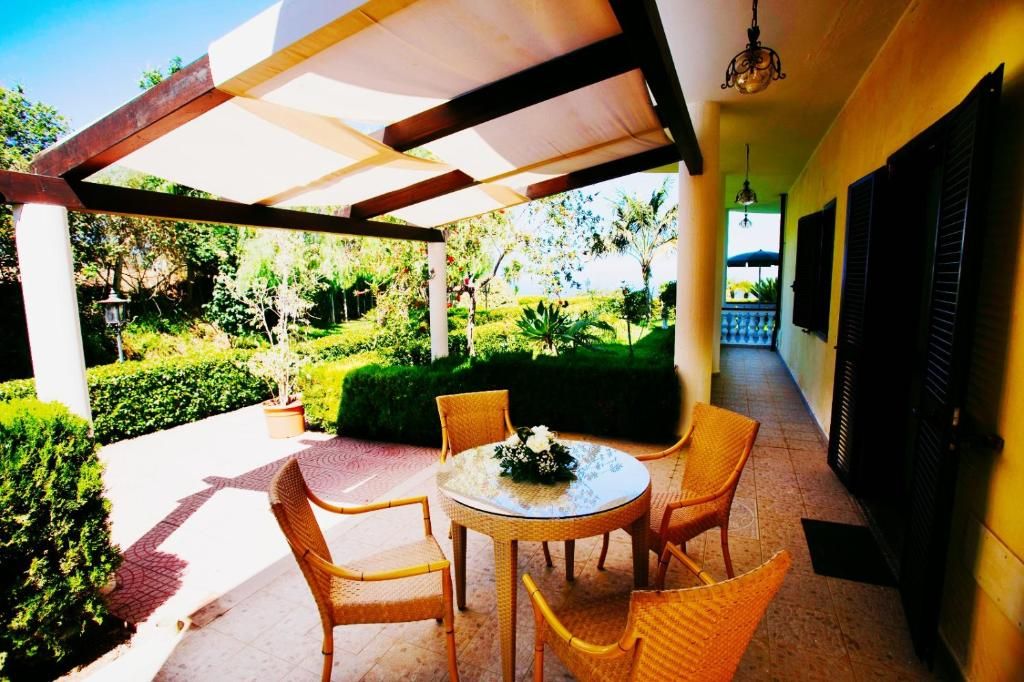 A covered outdoor seating area with wicker chairs and a table, surrounded by lush greenery and a clear view of the garden.