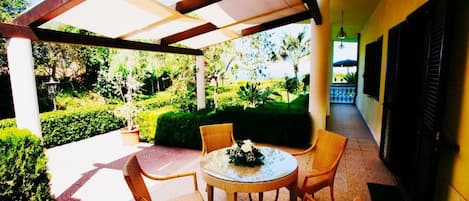 A covered outdoor seating area with wicker chairs and a table, surrounded by lush greenery and a clear view of the garden.