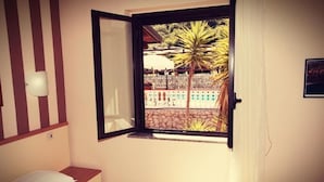Et soveværelse med en seng, et vindue der viser udsigt til en pool og palmer, samt en vægmonteret lampe.