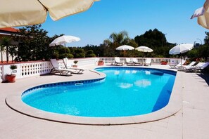 En swimmingpool med solstolar och parasoller, omgiven av ett vitt staket och grönska.