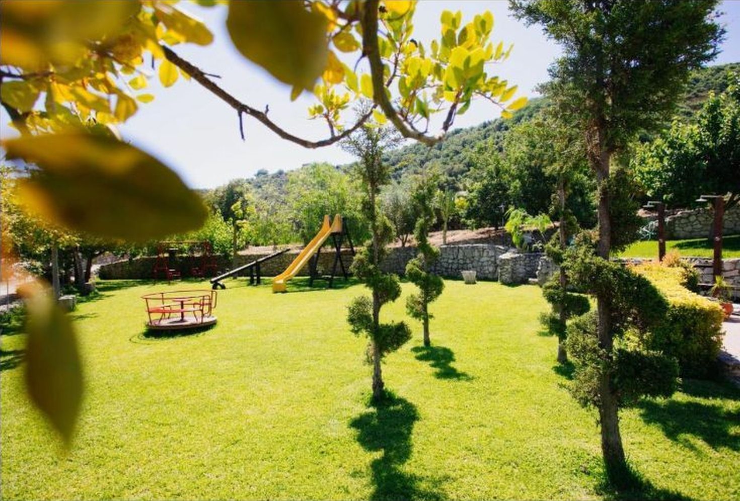 Un área verde bien cuidada con un parque infantil que incluye un tobogán y columpios, rodeada de árboles y un muro de piedra.