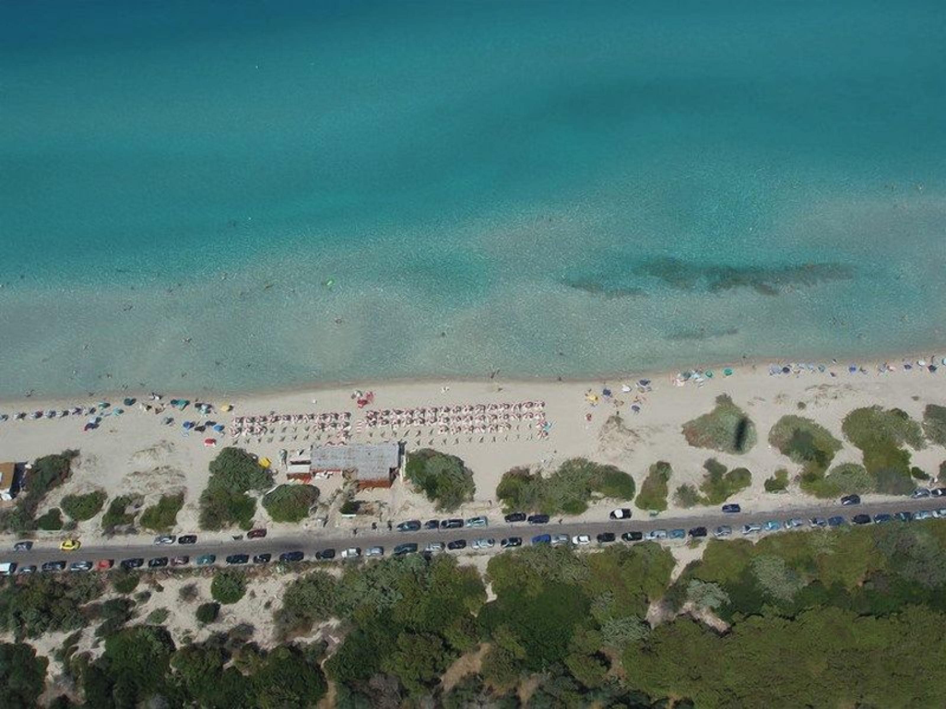 Vista aerea di una spiaggia con file di auto parcheggiate, una strada e un edificio con tetto rosso.