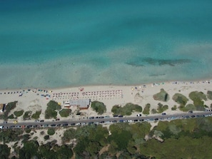 Une vue aérienne d’une plage où l’on aperçoit une rangée de voitures garées, une route et un bâtiment avec un toit rouge.