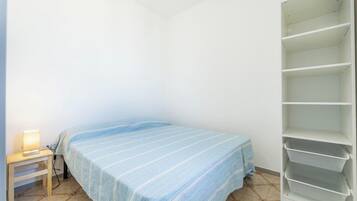 Ein kleines, einfach möbliertes Zimmer mit einem Bett, einem Nachttisch und einer weißen Regaleinheit.