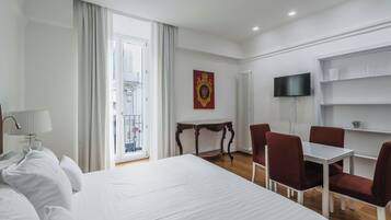 Una camera d'albergo con un letto grande, un tavolo da pranzo con sedie, una TV a parete e una finestra con tende.
