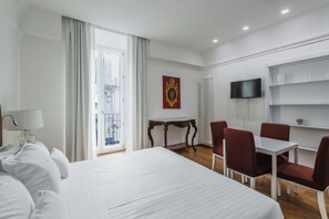 A hotel room with a large bed, a dining table with chairs, a TV mounted on the wall, and a window with curtains.