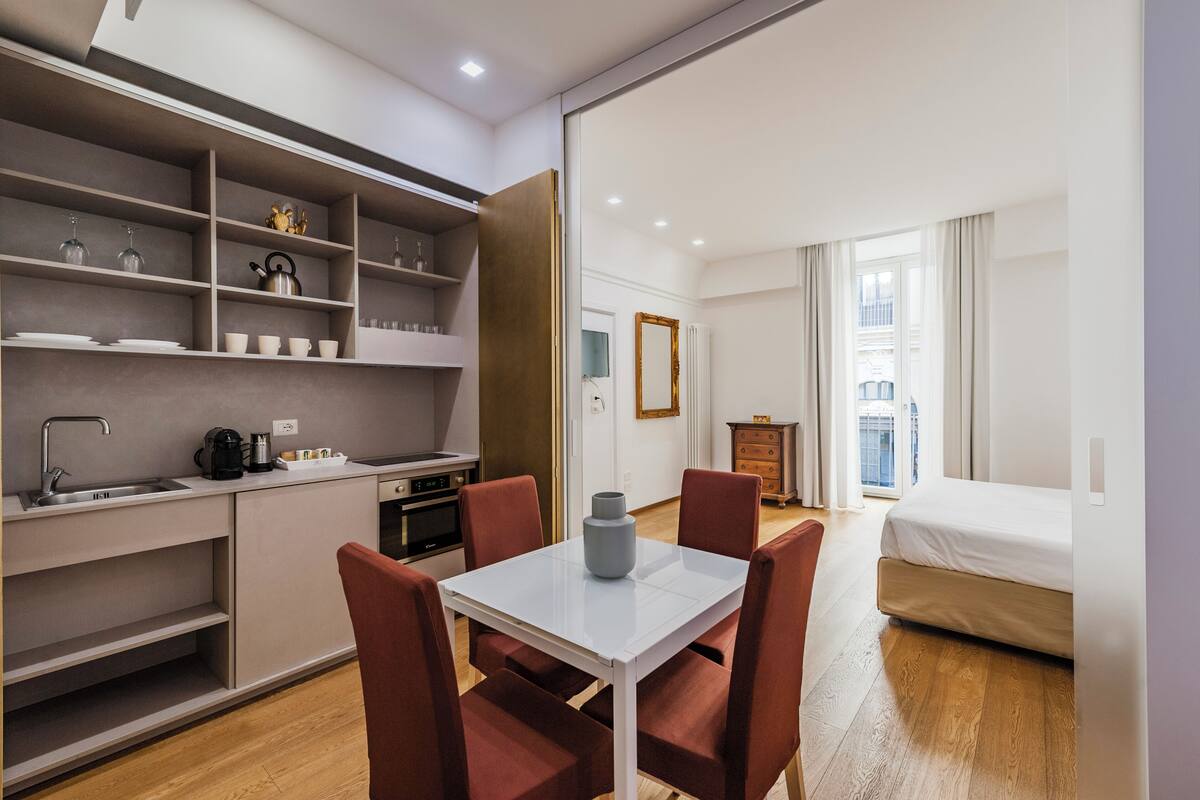 Habitación de hotel compacta con kitchenette, comedor con mesa y sillas, cama y vistas a la ciudad a través de amplias ventanas.
