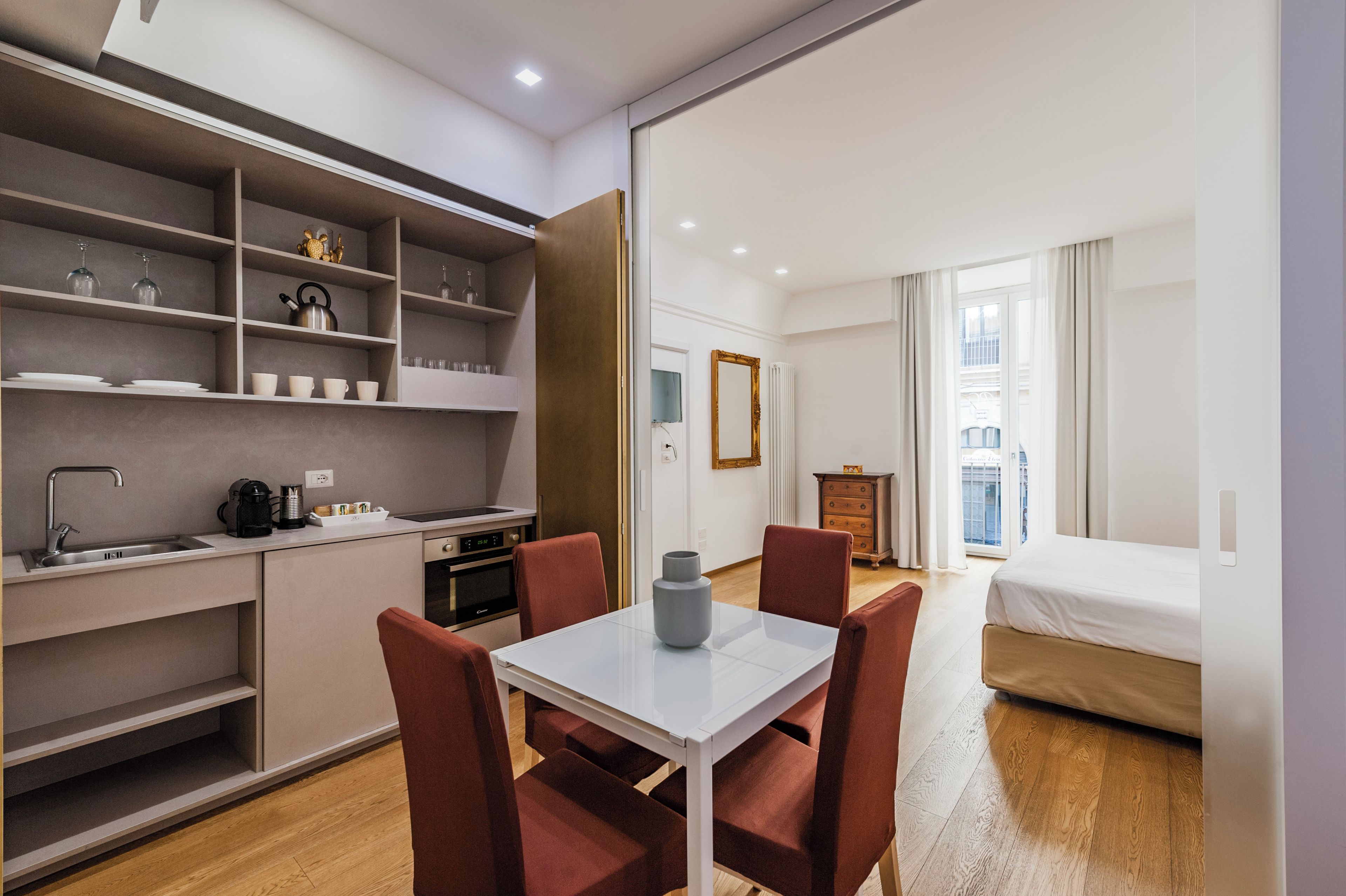 Una camera d'albergo compatta con angolo cottura, zona pranzo con tavolo e sedie, un letto e vista sulla città attraverso ampie finestre.