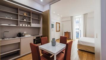 Una camera d'albergo compatta con angolo cottura, zona pranzo con tavolo e sedie, un letto e vista sulla città attraverso ampie finestre.