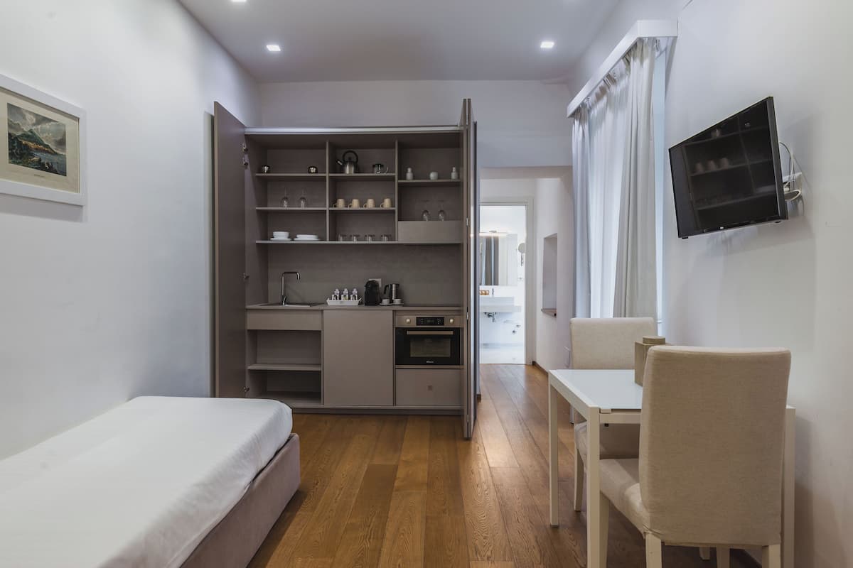 Habitación compacta con cama, escritorio, silla, televisión montada en la pared y kitchenette con estantes y electrodomésticos.