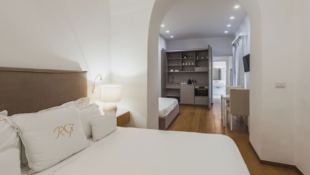 A hotel room with a bed, a nightstand, a lamp, an arched doorway leading to a kitchenette, and a dining area. - Rg7 Apartments (Catania)