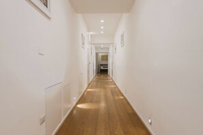 A narrow hallway with wooden flooring, white walls, and recessed lighting.