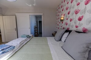 Ein Schlafzimmer mit einem Bett, Kissen, einem Nachttisch und einer Wandleuchte. Eine Tür führt in einen weiteren Raum.