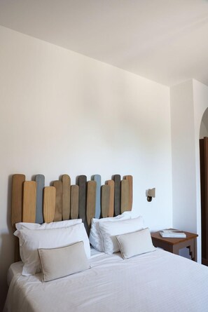 Una cama bien hecha con sábanas y almohadas blancas, un cabecero de madera con un diseño singular y una mesita de noche con un libro.
