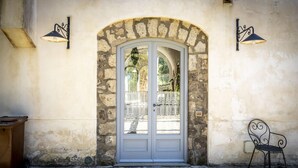Un bâtiment en pierre avec une porte blanche en arc de cercle, deux lampes fixées au mur et une chaise en métal à l’extérieur.