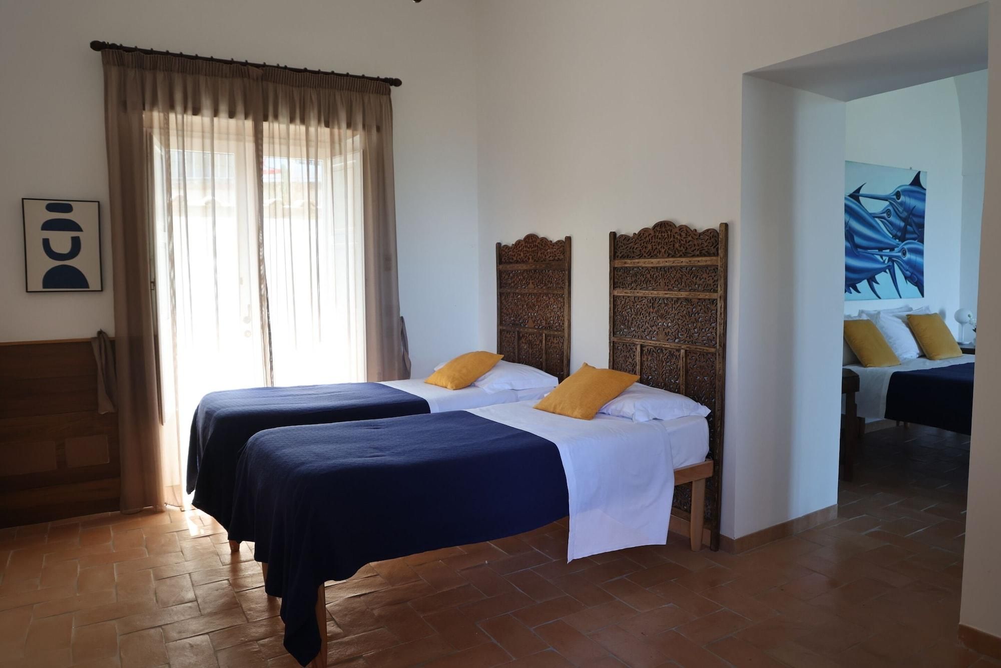 Une chambre d’hôtel avec deux lits, une grande fenêtre avec des rideaux légers, une tête de lit en bois et un tableau au mur.