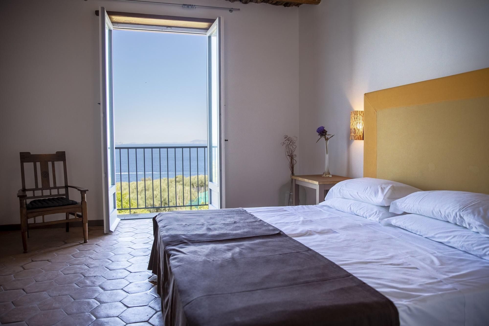 A bedroom with a bed, a chair, a nightstand, a lamp, and a view of the sea through a balcony door.