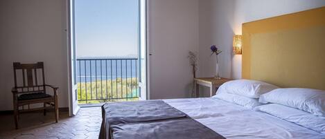 A bedroom with a bed, a chair, a nightstand, a lamp, and a view of the sea through a balcony door.