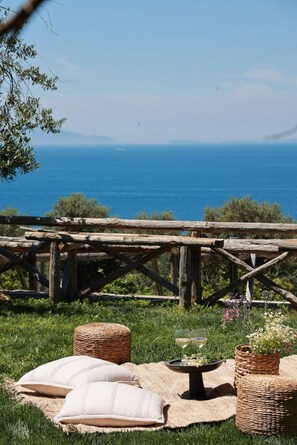 Un espacio de estar al aire libre rústico con cestas de mimbre, cojines y una mesa dispuesta para una comida, con vista al mar azul y despejado.