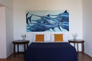 A bedroom with a large painting of fish on the wall, two bedside tables with lamps, and a bed with white and mustard pillows.