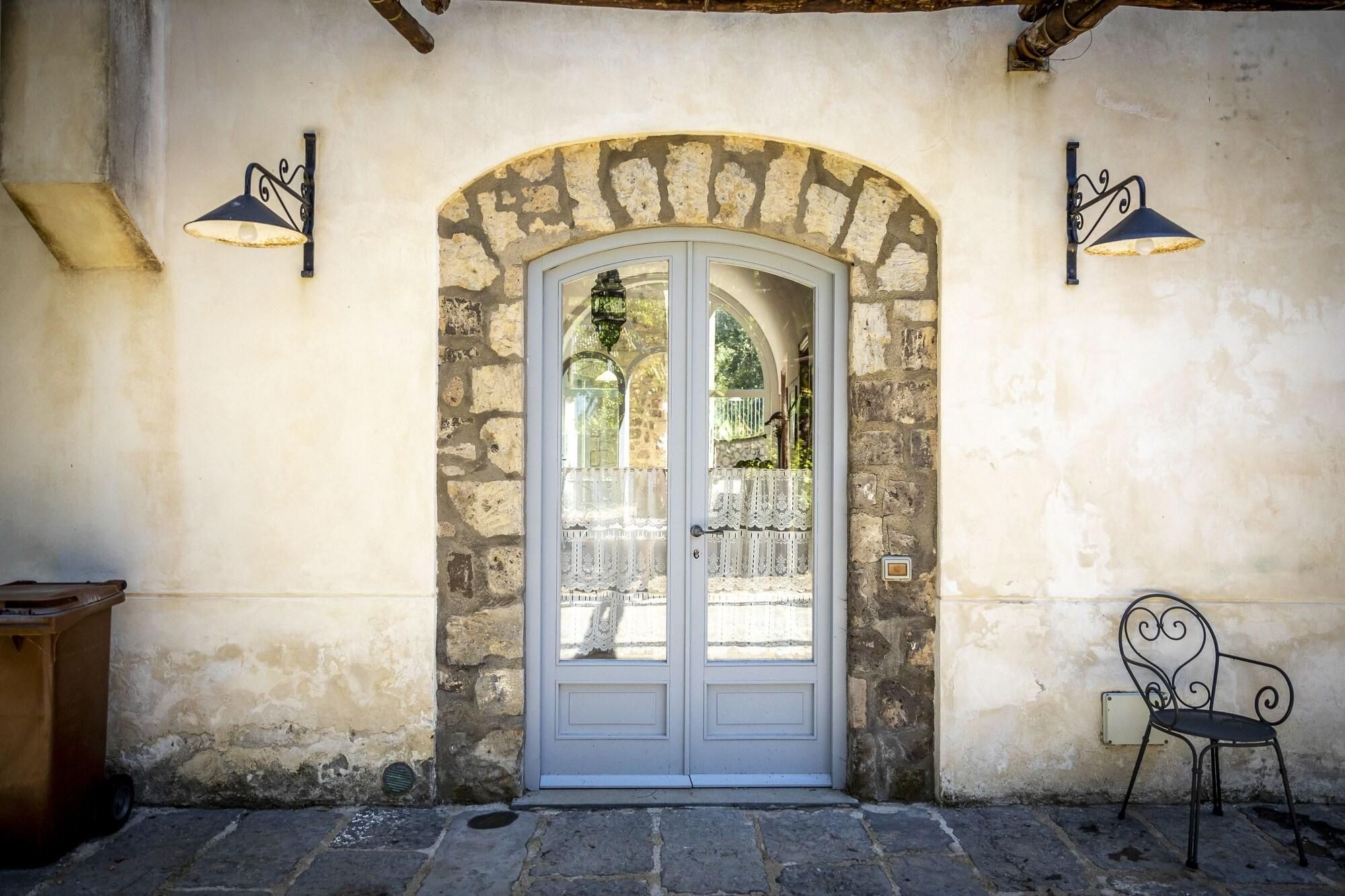 Un edificio in pietra con una porta ad arco bianca, due lampioni a muro e una sedia di metallo all'esterno.