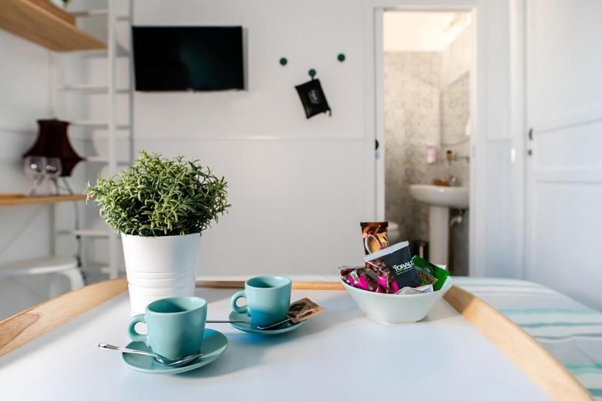 A neatly arranged breakfast tray with a potted plant, two teal cups, and a bowl of assorted pastries on a white table in a hotel room.