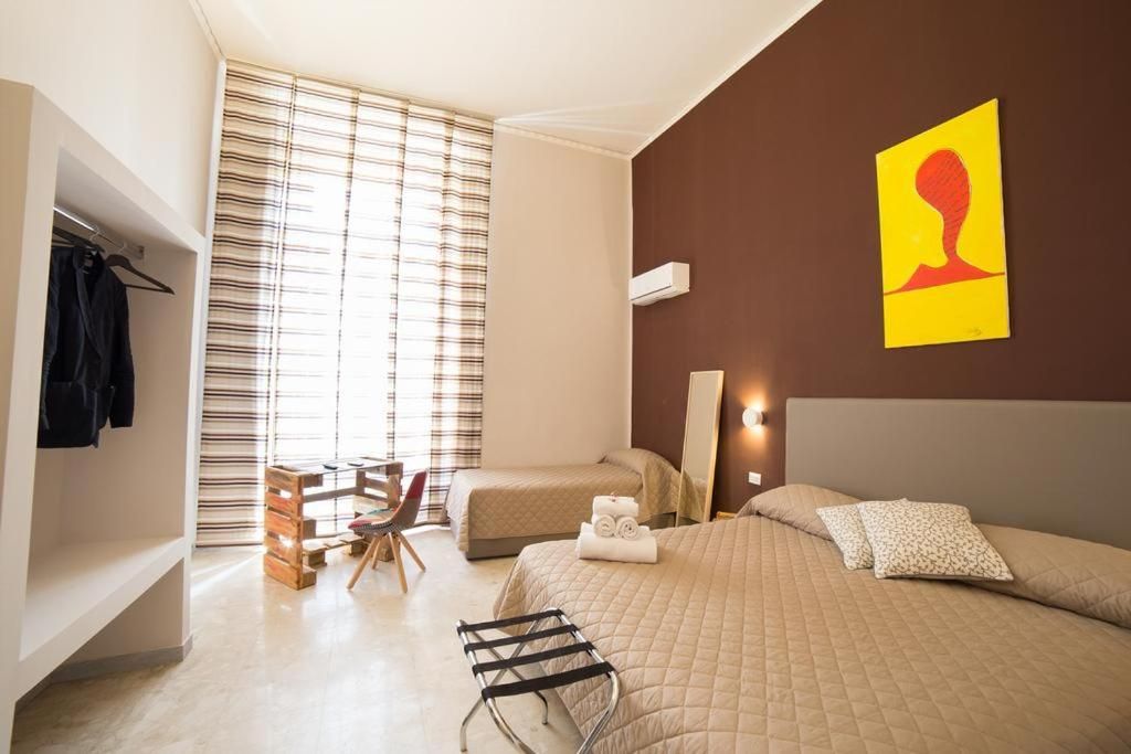 A modern bedroom with a large bed, a desk, and a chair. There is a window with blinds, a coat rack, and a painting on the wall.