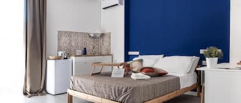 A modern hotel room with a bed, bedside table, and a small kitchenette.