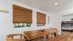 A room with a wooden dining table, two chairs, a bunk bed, and large windows with blinds.