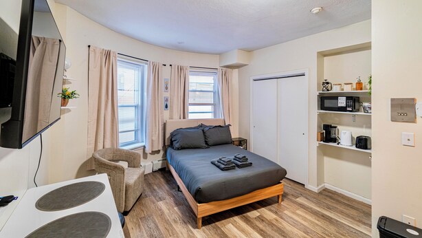 Una habitación pequeña con cama, escritorio, silla, televisor y una zona de kitchenette.