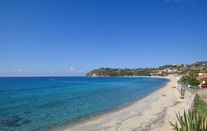 Una playa costera con aguas azules transparentes, una orilla arenosa y una colina con edificios al fondo.